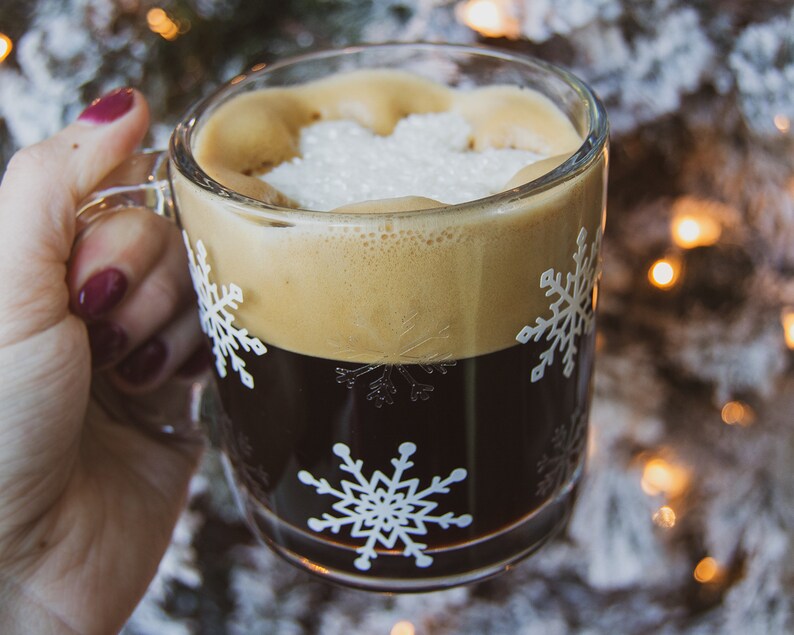 May include: A clear glass mug filled with dark coffee and topped with foam and sugar. The mug features white snowflake designs. A hand with red nail polish holds the mug. The background is blurred with Christmas lights.