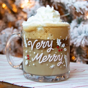 Puede incluir: Una taza de vidrio transparente con asa blanca llena de una bebida de caf&eacute; marr&oacute;n coronada con crema batida. La taza tiene un dise&ntilde;o blanco que dice "Very Merry" con acebo y copos de nieve.