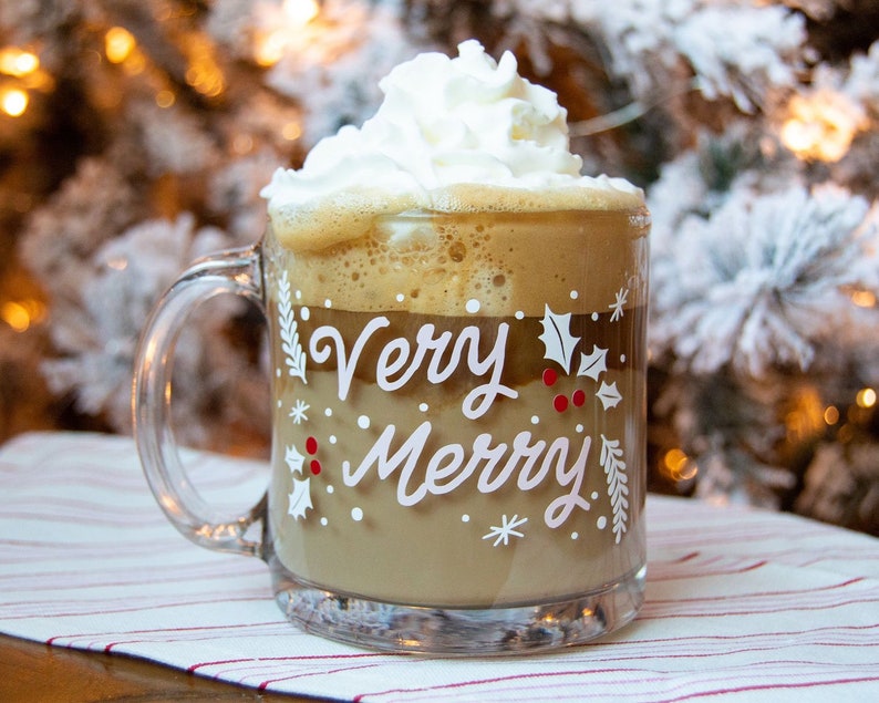 Puede incluir: Una taza de vidrio transparente con asa blanca llena de una bebida de caf&eacute; marr&oacute;n coronada con crema batida. La taza tiene un dise&ntilde;o blanco que dice "Very Merry" con acebo y copos de nieve.