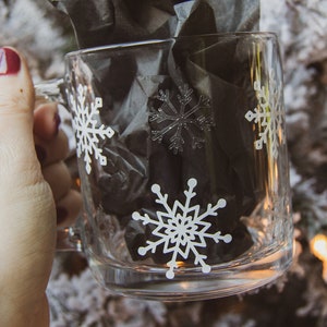 May include: A clear glass mug decorated with white snowflakes. The mug contains black tissue paper. The mug is held by a person with dark red nail polish. The background is a blurred image of a snowy Christmas tree.