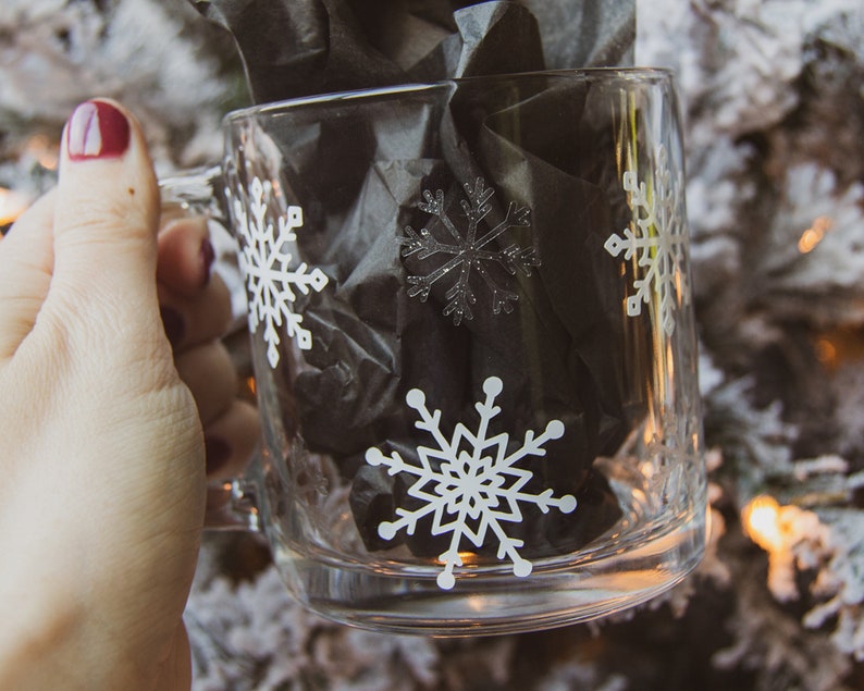 May include: A clear glass mug decorated with white snowflakes. The mug contains black tissue paper. The mug is held by a person with dark red nail polish. The background is a blurred image of a snowy Christmas tree.