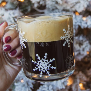 May include: A clear glass mug filled with dark coffee and topped with foamy cream. The mug features white snowflake designs. The mug is held by a person with red nail polish. The background is blurred.