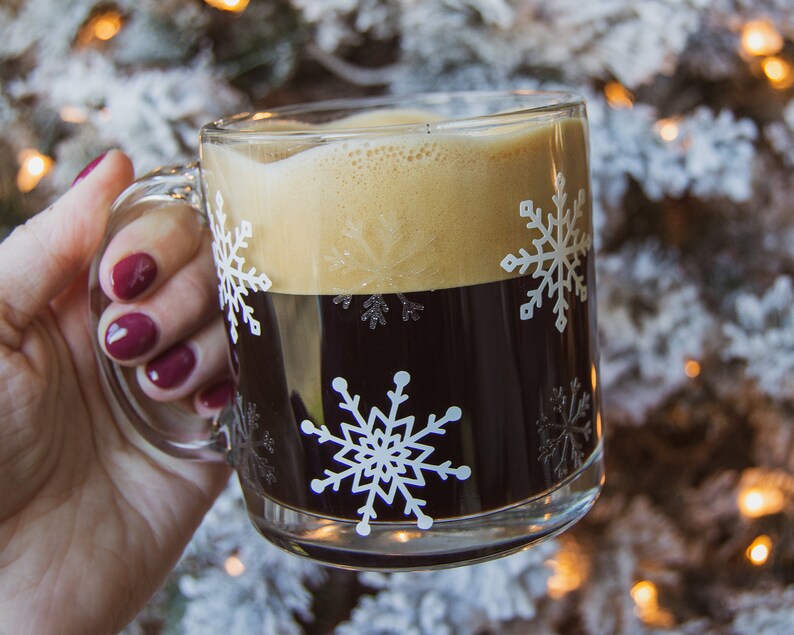May include: A clear glass mug filled with dark coffee and topped with foamy cream. The mug features white snowflake designs. The mug is held by a person with red nail polish. The background is blurred.