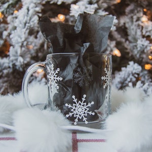 May include: A clear glass mug with a handle, adorned with white snowflake patterns. The mug is filled with black tissue paper and rests on a white and red plaid surface, with a blurred Christmas tree in the background.
