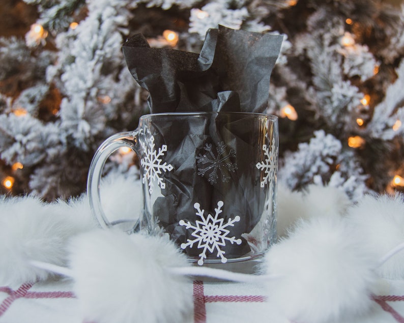 May include: A clear glass mug with a handle, adorned with white snowflake patterns. The mug is filled with black tissue paper and rests on a white and red plaid surface, with a blurred Christmas tree in the background.