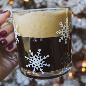 May include: A clear glass mug filled with dark coffee and topped with a layer of tan foam. The mug is decorated with white snowflake designs. The background is a blurred image of a Christmas tree with lights.