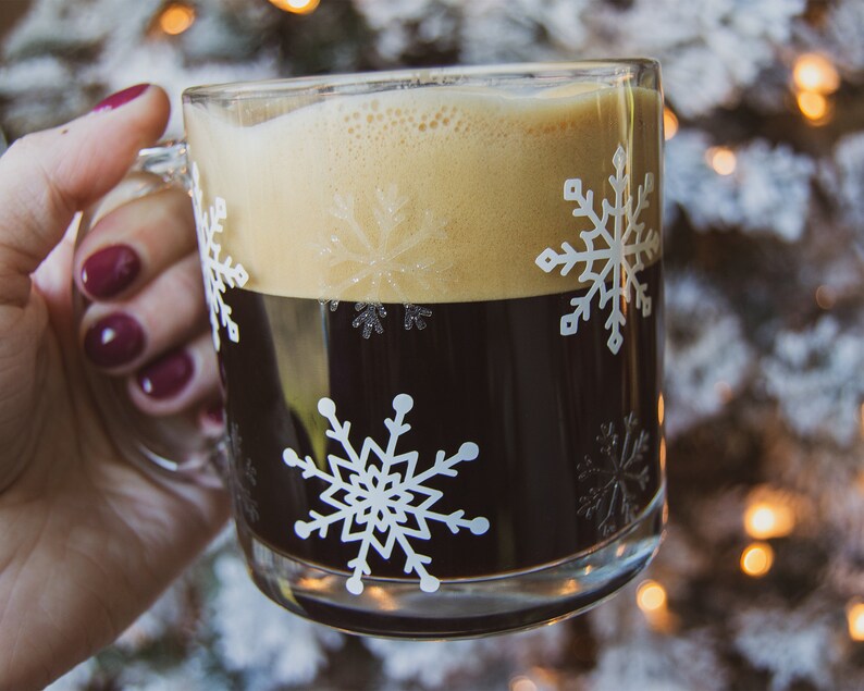 May include: A clear glass mug filled with dark coffee and topped with a layer of tan foam. The mug is decorated with white snowflake designs. The background is a blurred image of a Christmas tree with lights.