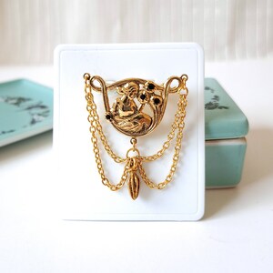 May include: A gold-toned brooch featuring a central circular relief of a seated figure surrounded by three small black stones. The piece includes two draped gold chains connecting to a central dangling pendant, all mounted on a white display card.
