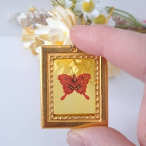 May include: A gold-toned pendant featuring a framed image of a red butterfly with black markings. The pendant is held by a finger against a white background.