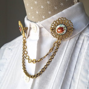 May include: A gold-tone metal collar pin set featuring two circular brooches with intricate filigree edges and a central floral motif painted in red and green. The two brooches are connected by two draped gold-tone metal chains of varying lengths.