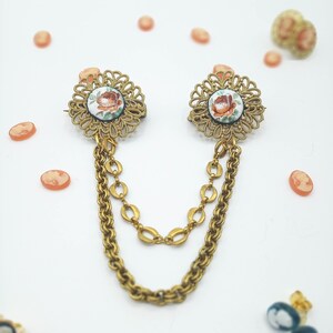 May include: A gold-toned metal sweater clip featuring two circular filigree brooches connected by two draped chains. Each brooch contains a central white disc with a painted orange and green floral motif, surrounded by small orange cameo accents.