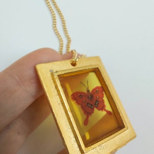 May include: A gold chain necklace with a pendant featuring a red butterfly encased in a clear resin frame. The frame is surrounded by a gold border.