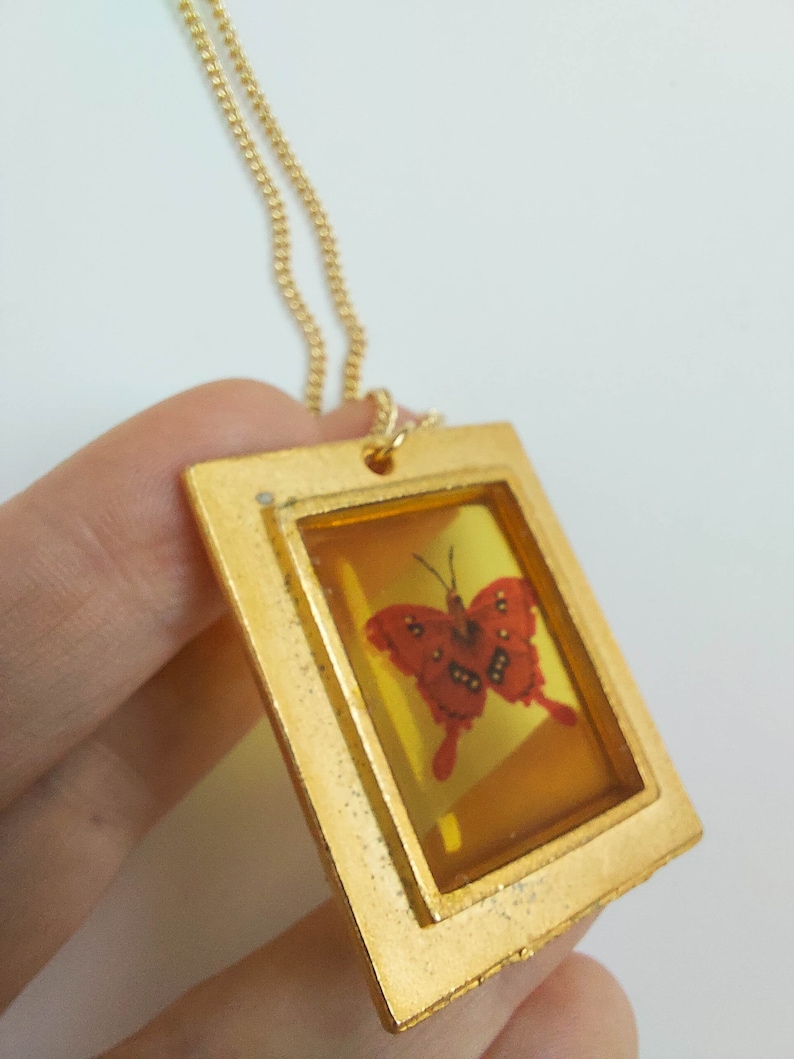 May include: A gold chain necklace with a pendant featuring a red butterfly encased in a clear resin frame. The frame is surrounded by a gold border.