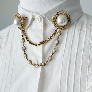 May include: A gold-toned collar brooch featuring two circular filigree settings, each centered with a large faux pearl. The two pieces are connected by a double-strand chain, with one chain featuring small inset faux pearls along its length.