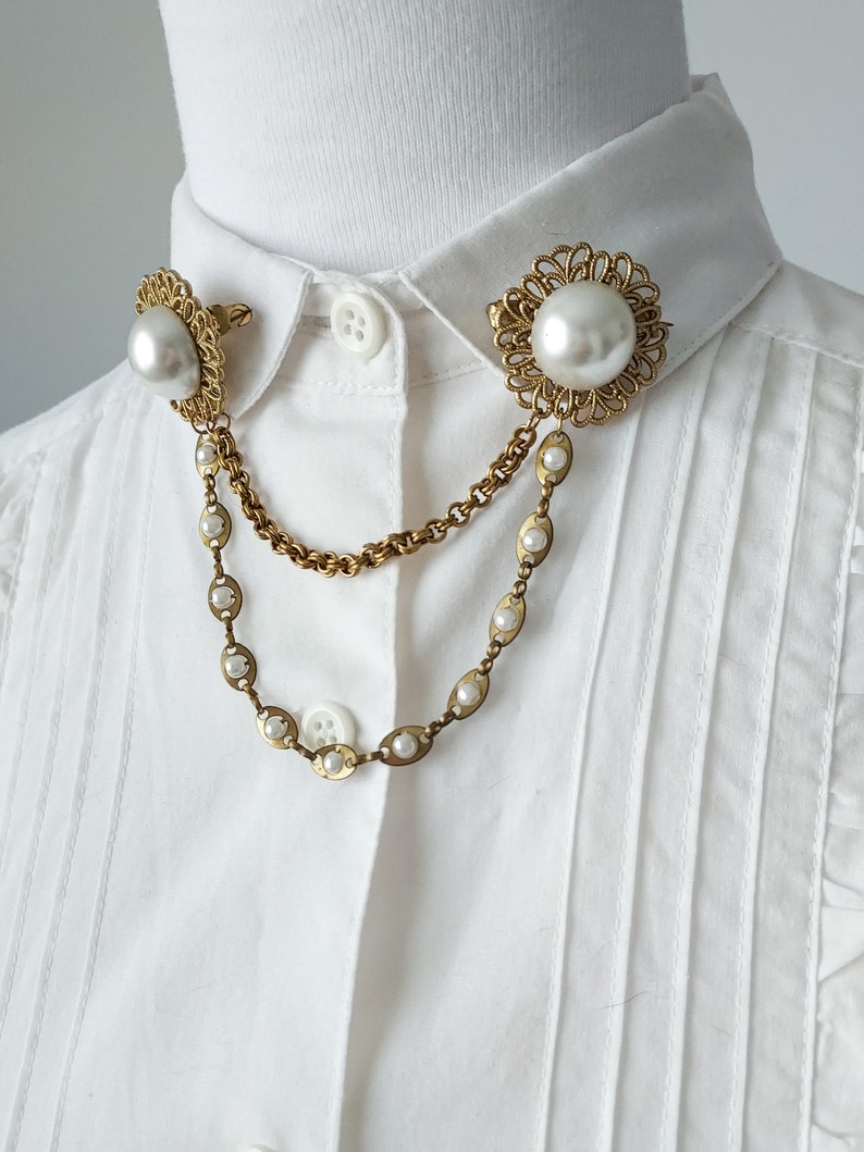 May include: A gold-toned collar brooch featuring two circular filigree settings, each centered with a large faux pearl. The two pieces are connected by a double-strand chain, with one chain featuring small inset faux pearls along its length.
