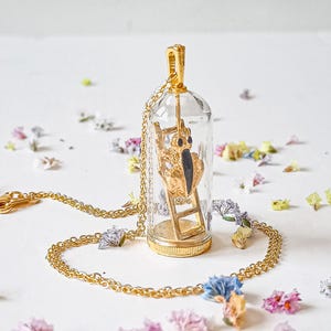 May include: A gold necklace with a glass pendant featuring a small golden figure climbing a ladder. The pendant is clear and the figure is visible inside.