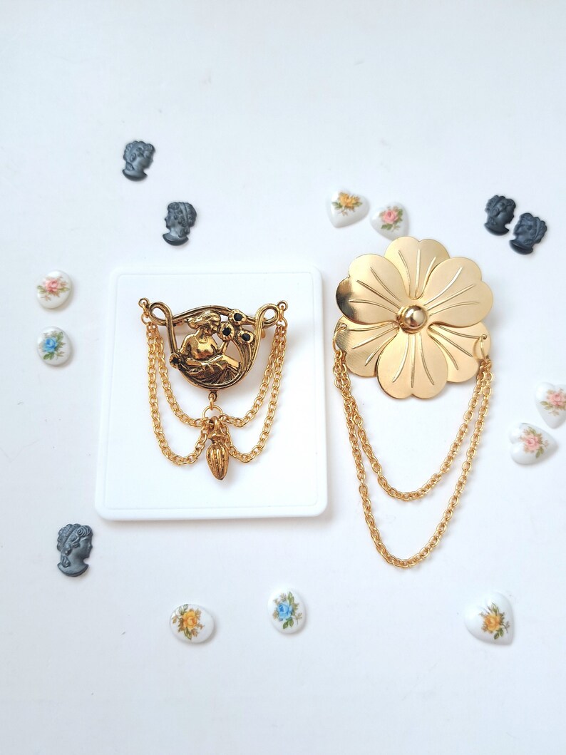 May include: Two gold-tone metal brooches. One brooch features a central figure with two dangling chains and a small pendant. The other brooch is a large, six-petalled flower with two long, draped gold chains. Small decorative cabochons surround them.