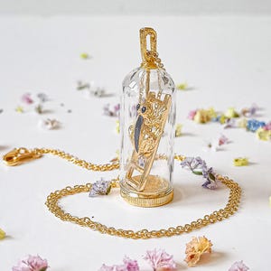 May include: A gold necklace with a glass pendant featuring a small, black and gold figurine climbing a ladder. The pendant is suspended from a gold chain.
