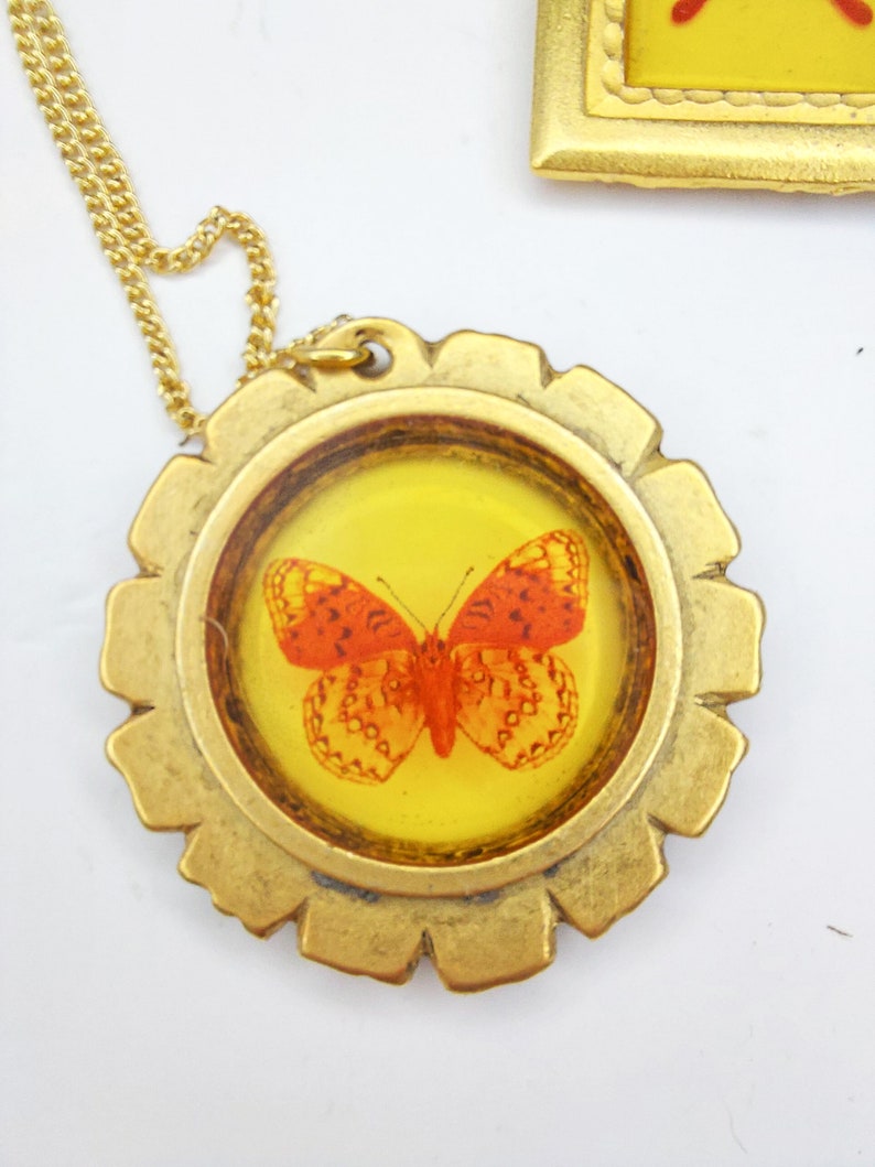May include: Gold necklace with a round pendant featuring a framed orange butterfly with black markings. The pendant has a textured, gear-like design.