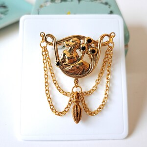 May include: A gold-tone brooch featuring a central circular medallion with a relief figure of a person holding flowers, accented with small black stones. The piece includes two draped gold chains and a central dangling teardrop-shaped charm.