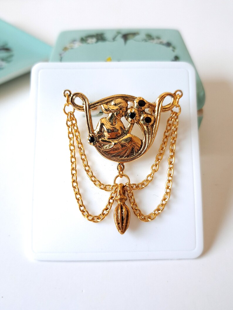 May include: A gold-tone brooch featuring a central circular medallion with a relief figure of a person holding flowers, accented with small black stones. The piece includes two draped gold chains and a central dangling teardrop-shaped charm.