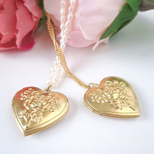 Op de afbeelding: Twee gouden hartvormige hangers met een roosontwerp hangen aan een parel- en gouden ketting. De hangers bevinden zich op een witte achtergrond.