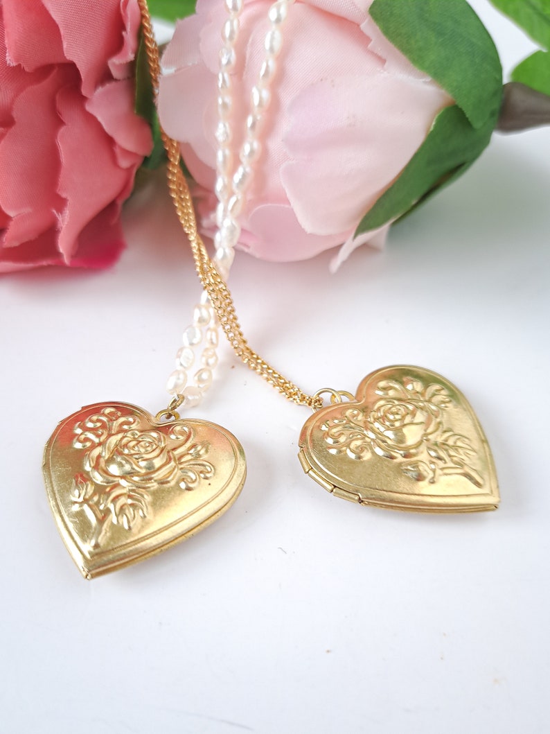 Op de afbeelding: Twee gouden hartvormige hangers met een roosontwerp hangen aan een parel- en gouden ketting. De hangers bevinden zich op een witte achtergrond.