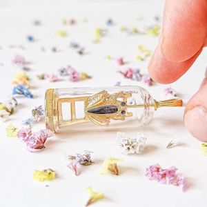 May include: A gold-colored frog figurine is suspended inside a clear glass bottle. The frog is perched on a miniature gold ladder. The bottle is lying on a white surface with scattered flower petals.