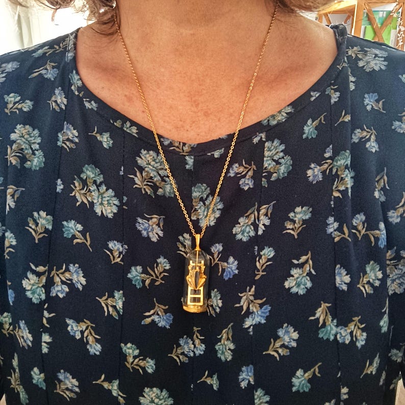 May include: A gold chain necklace with a pendant featuring a small, clear glass vial containing a miniature golden ladder. The necklace is worn by a person with a dark blue floral patterned shirt.