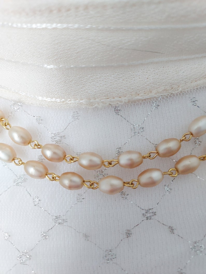 May include: Two strands of a pearl necklace with gold chain links. The pearls are a pale pink color and are oval shaped.