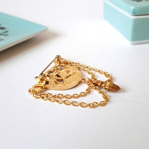 May include: A gold-toned metal brooch featuring a circular central medallion with an embossed design. Two delicate gold chains drape from the medallion, connecting to a small, textured, pointed charm. A pin mechanism is attached to the back.