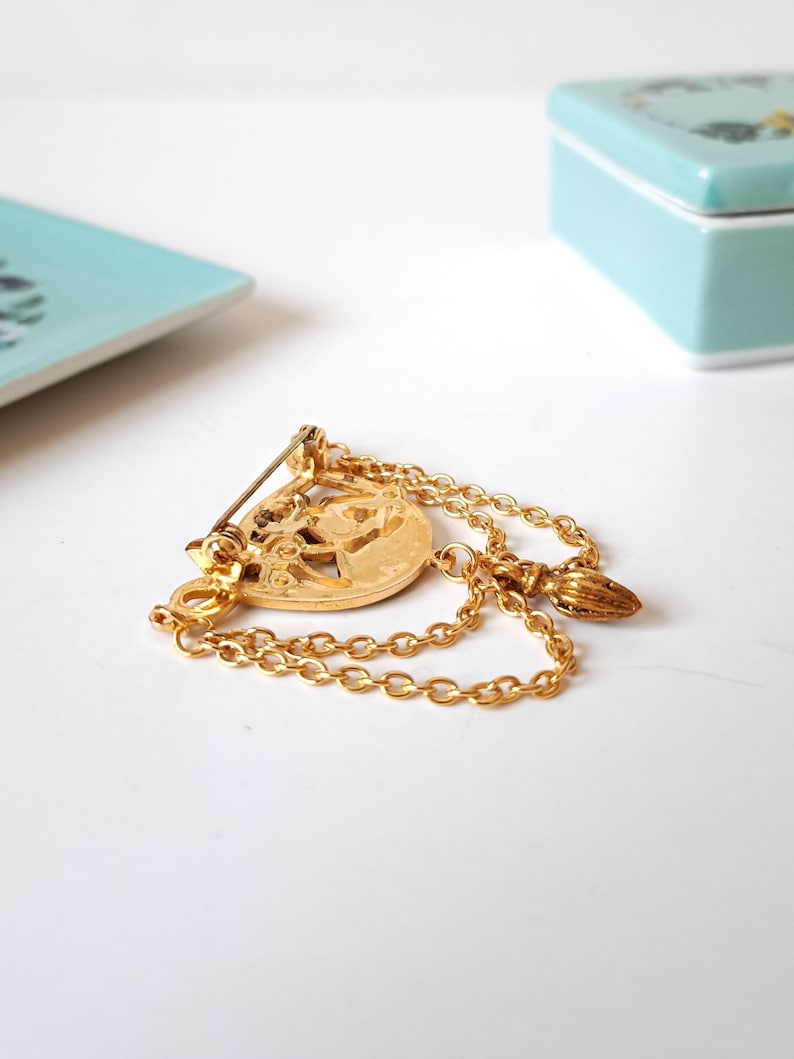 May include: A gold-toned metal brooch featuring a circular central medallion with an embossed design. Two delicate gold chains drape from the medallion, connecting to a small, textured, pointed charm. A pin mechanism is attached to the back.