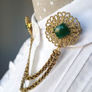 May include: A gold-tone metal collar pin featuring two circular filigree brooches connected by a double chain. Each brooch is centered with a square, dark green stone. The accessory is attached to the collar of a white, button-down shirt.