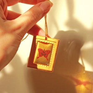 May include: A gold chain necklace with a gold frame pendant featuring a red butterfly inside. The pendant is rectangular and has a decorative border.