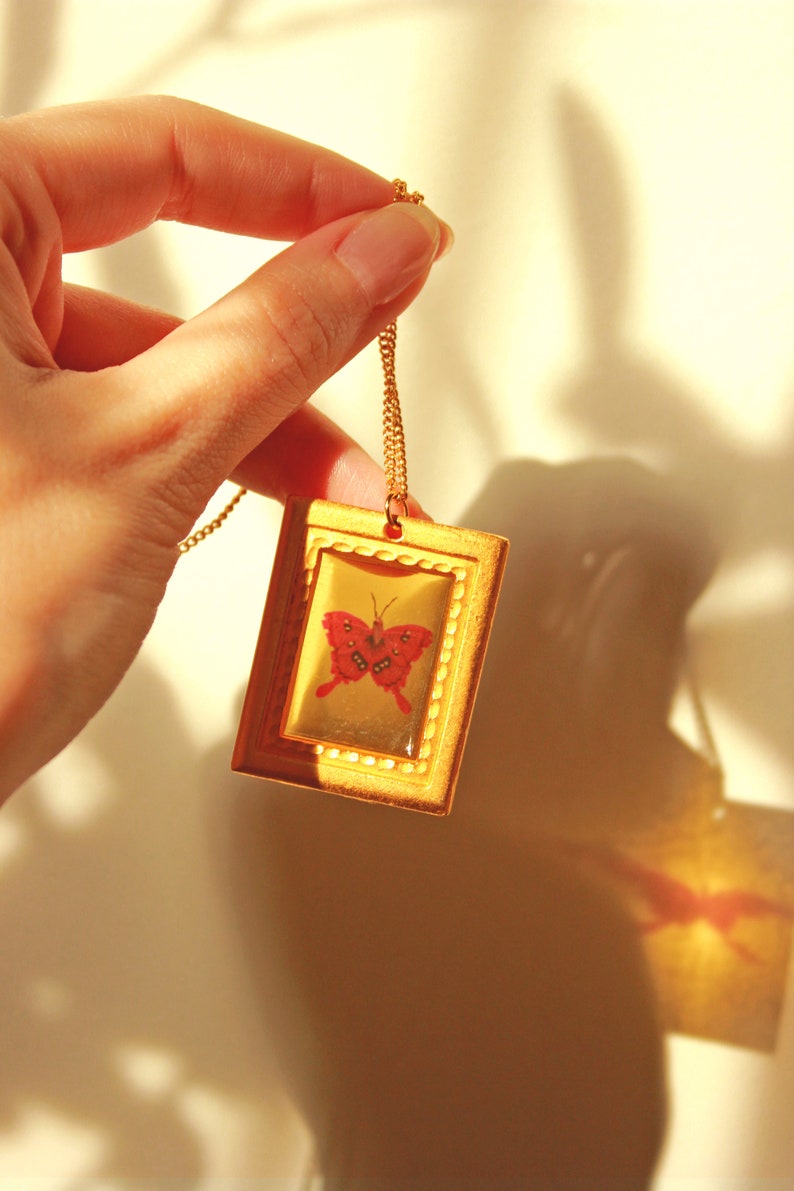May include: A gold chain necklace with a gold frame pendant featuring a red butterfly inside. The pendant is rectangular and has a decorative border.