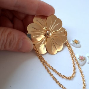 May include: A gold-toned metal brooch shaped like a five-petaled flower with a smooth, polished finish. The centre features a small, raised gold sphere. Two delicate gold-toned metal chains are attached to the bottom petals, creating a draped effect.