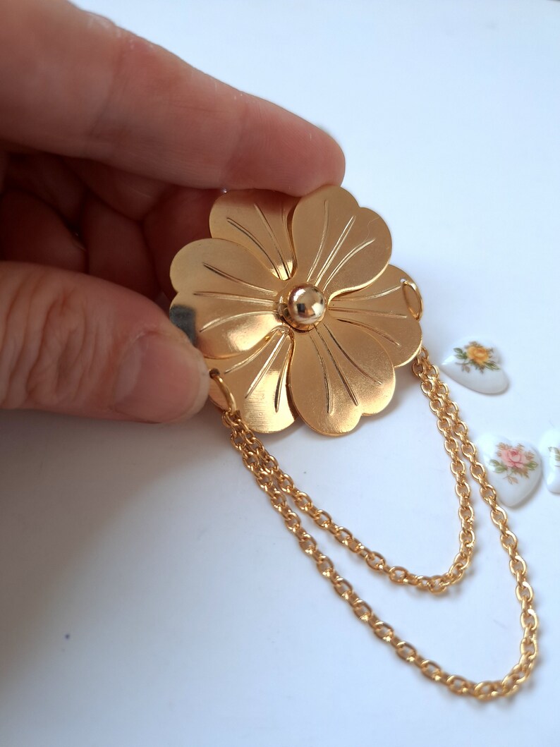 May include: A gold-toned metal brooch shaped like a five-petaled flower with a smooth, polished finish. The centre features a small, raised gold sphere. Two delicate gold-toned metal chains are attached to the bottom petals, creating a draped effect.