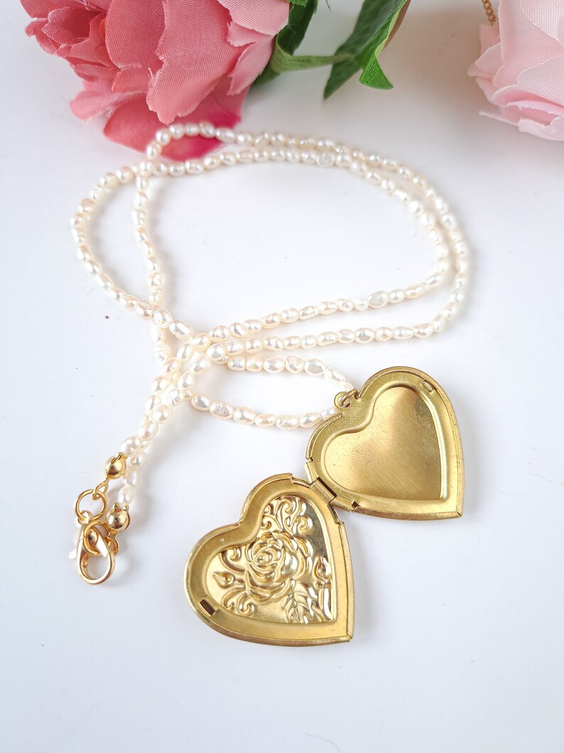 Op de afbeelding: Een gouden hartvormige hanger met een bloemenmotief aan de ene kant en een glad oppervlak aan de andere kant. De hanger is bevestigd aan een witte parelketting.