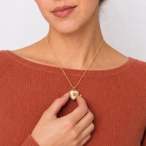 May include: A gold-toned necklace featuring a heart-shaped pendant with a white center and a small pink floral design. The pendant hangs from a delicate gold-toned chain, resting against a rust-colored ribbed knit top worn by an individual.