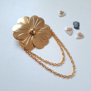 May include: A gold-toned metal brooch shaped like a five-petaled flower with a smooth, polished finish. The centre features a small, raised gold sphere. Two delicate gold-toned link chains are attached to the flower, creating a draped effect.