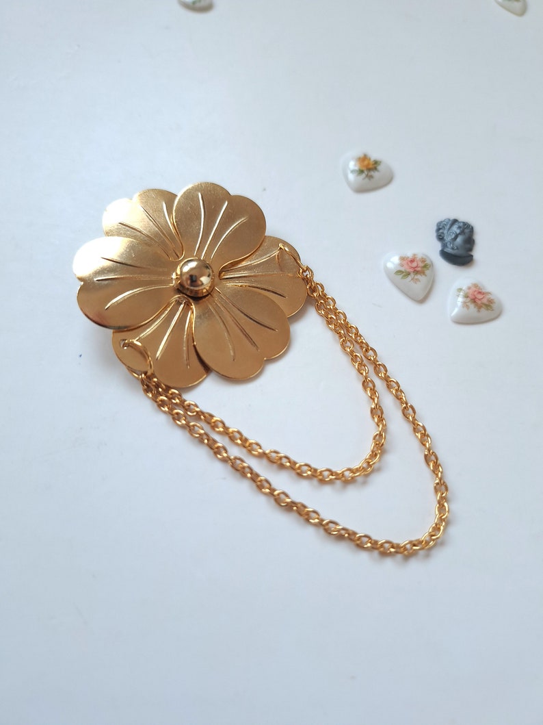 May include: A gold-toned metal brooch shaped like a five-petaled flower with a smooth, polished finish. The centre features a small, raised gold sphere. Two delicate gold-toned link chains are attached to the flower, creating a draped effect.