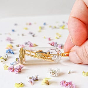May include: A gold-colored grasshopper figurine is encased in a clear glass vial with a gold-colored cap. The vial is lying on a white surface with scattered small, colorful flowers.