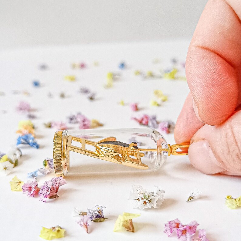 May include: A gold-colored grasshopper figurine is encased in a clear glass vial with a gold-colored cap. The vial is lying on a white surface with scattered small, colorful flowers.