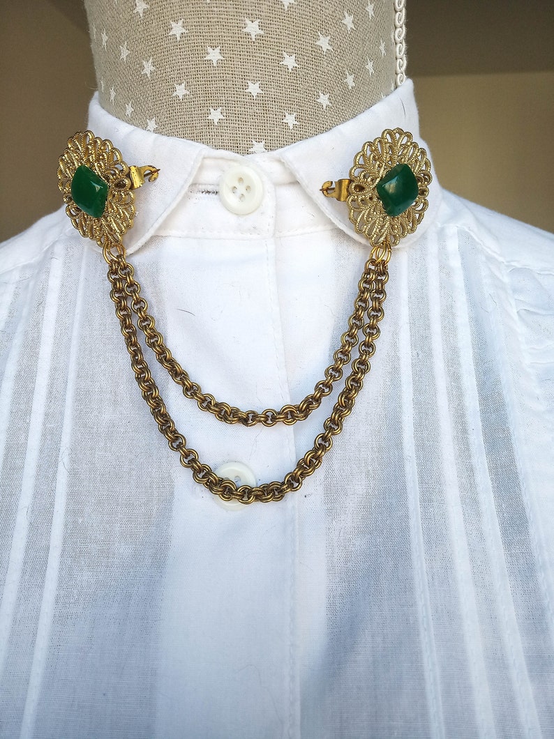 May include: A gold-tone metal collar clip featuring two ornate, filigree-style circular brooches connected by a double-strand chain. Each brooch is centered with a square, deep green stone. The accessory is attached to the collar of a white shirt.