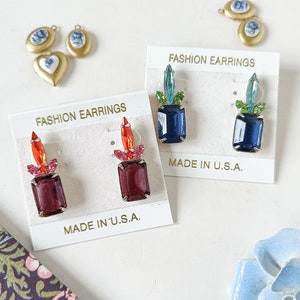 Pode incluir: Dois pares de brincos vintage em cartões brancos. Os brincos são feitos de pedras de vidro azul, verde, vermelho e roxo com detalhes dourados. Os cartões têm o texto "Fashion Earrings" e "Made in U.S.A."