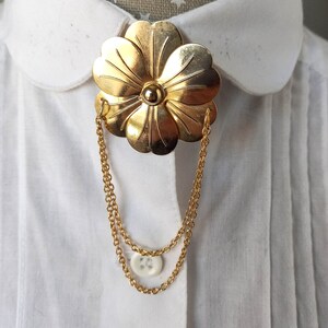 May include: A gold-toned metal brooch shaped like a five-petaled flower with a central sphere. Two delicate gold-toned chains hang from the sides of the flower, looping downward to connect to a small, round, white four-hole button.