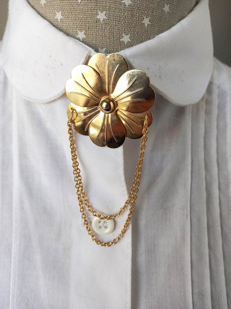 May include: A gold-toned metal brooch shaped like a five-petaled flower with a central sphere. Two delicate gold-toned chains hang from the sides of the flower, looping downward to connect to a small, round, white four-hole button.