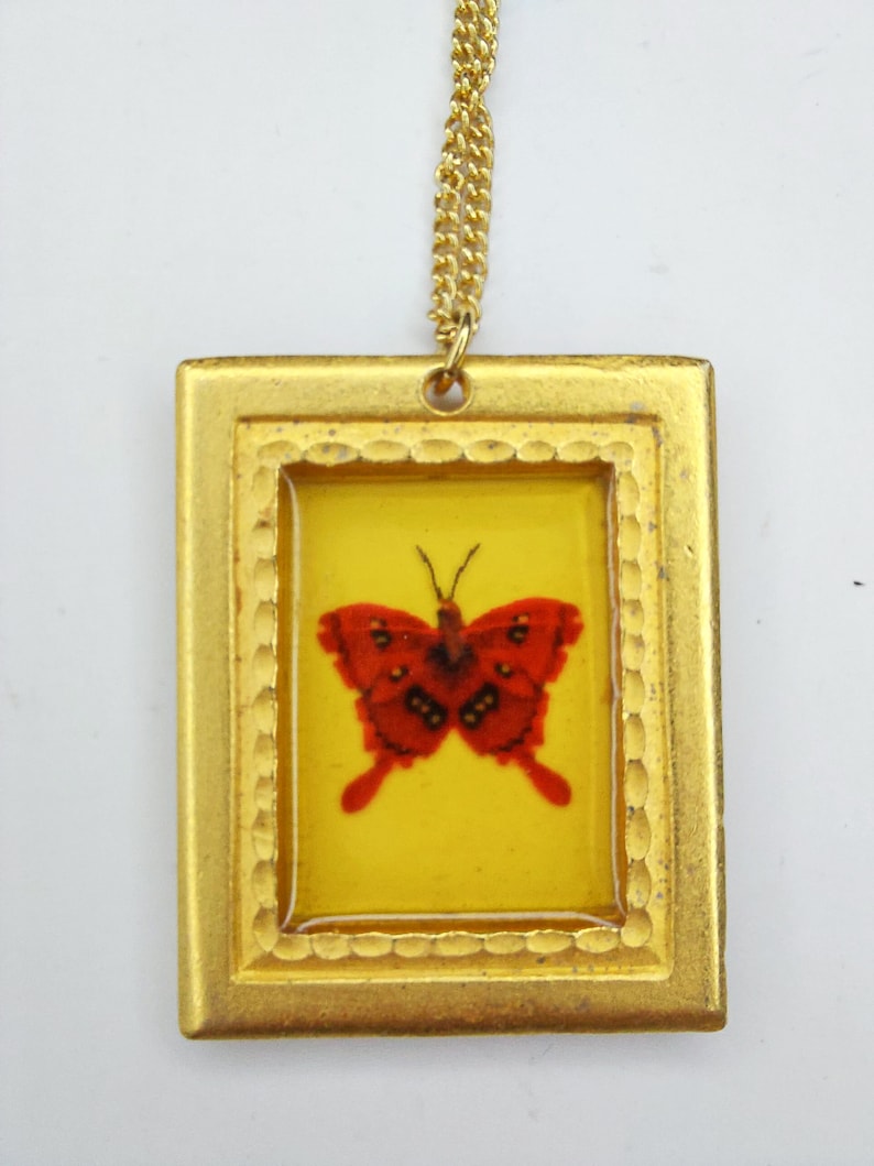 May include: A gold-toned pendant necklace with a red butterfly encased in a clear resin frame. The frame is surrounded by a gold-toned border with a textured design.
