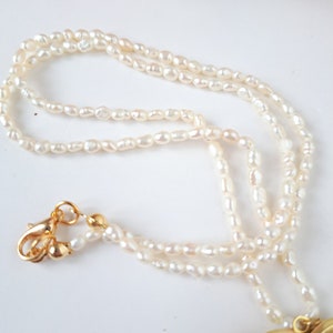 Op de afbeelding: Een witte parelketting met een gouden hartvormige hanger. De ketting is gemaakt van kleine, onregelmatige parels en heeft een gouden sluiting.
