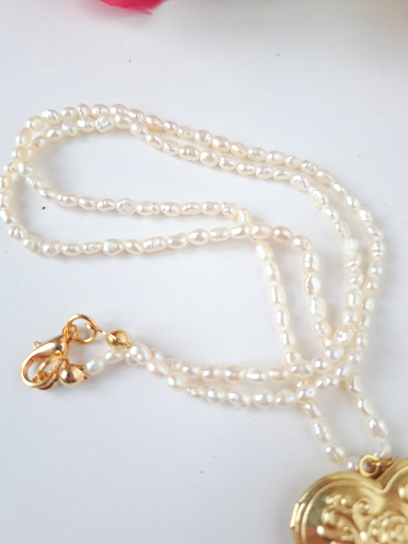 Op de afbeelding: Een witte parelketting met een gouden hartvormige hanger. De ketting is gemaakt van kleine, onregelmatige parels en heeft een gouden sluiting.